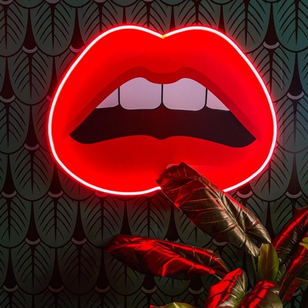 Vordergrund: eine Zimmerpflanze. Hintergrund und Fokus: eine große Grafik von einem offenen Mund, eingefasst von Neonlicht